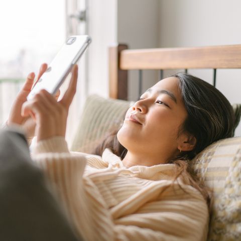 une jeune et belle femme est allongée dans son lit et utilise joyeusement un téléphone intelligent le matin