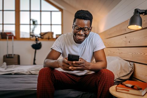 beau jeune homme souriant avec des lunettes dans des vêtements décontractés vérifiant son téléphone intelligent assis sur le lit